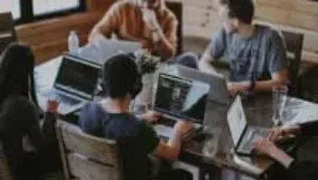four co-workers sitting at a desk looking at laptops