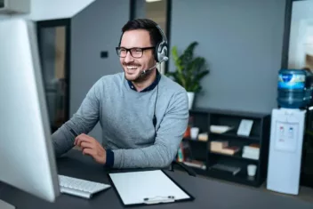 A man wearing headphones smiles widely whilst looking at his computer screen as he interacts with a person he interacts with
