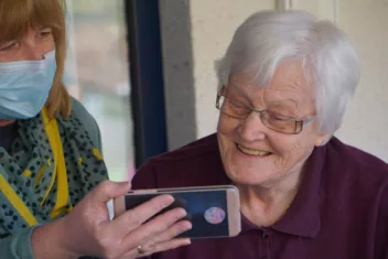 female Care home staff member in mask showing a phone to an smiling lady with short grey hair